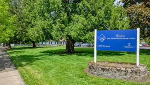 A blue sign for _Sapphire at Gresham Post-Acute Care and Rehabilitation_ on a stone base, surrounded by a large grassy lawn and trees