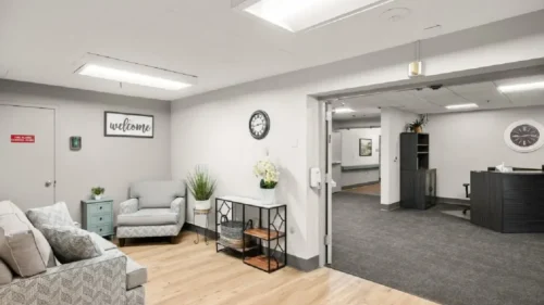 A common area with a patterned gray couch, a matching armchair, a side table, and a large open doorway leading to a reception area